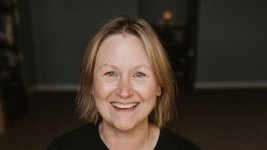 Blonde woman with a wide smile, wearing a black shirt, in front of a dark background.