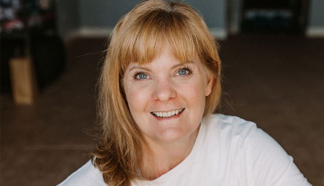 Woman with blonde hair and bangs smiles, wearing a white shirt, in a neutral-toned interior.