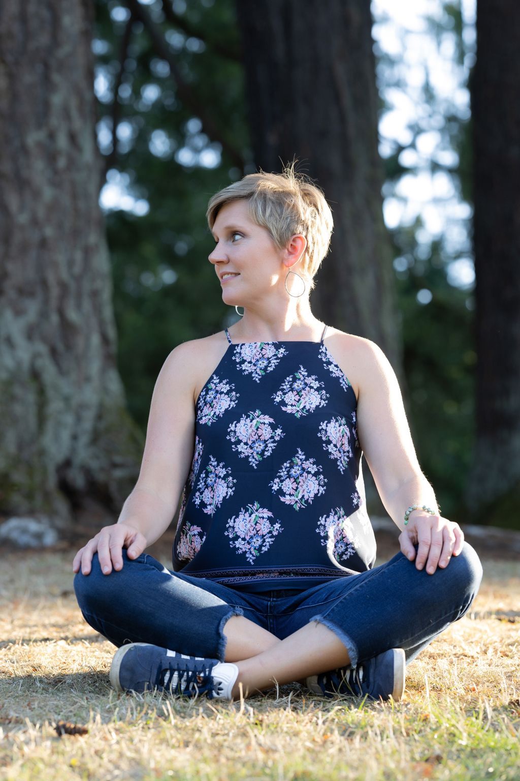 Woman in floral top sits cross-legged outdoors, looking to the side.