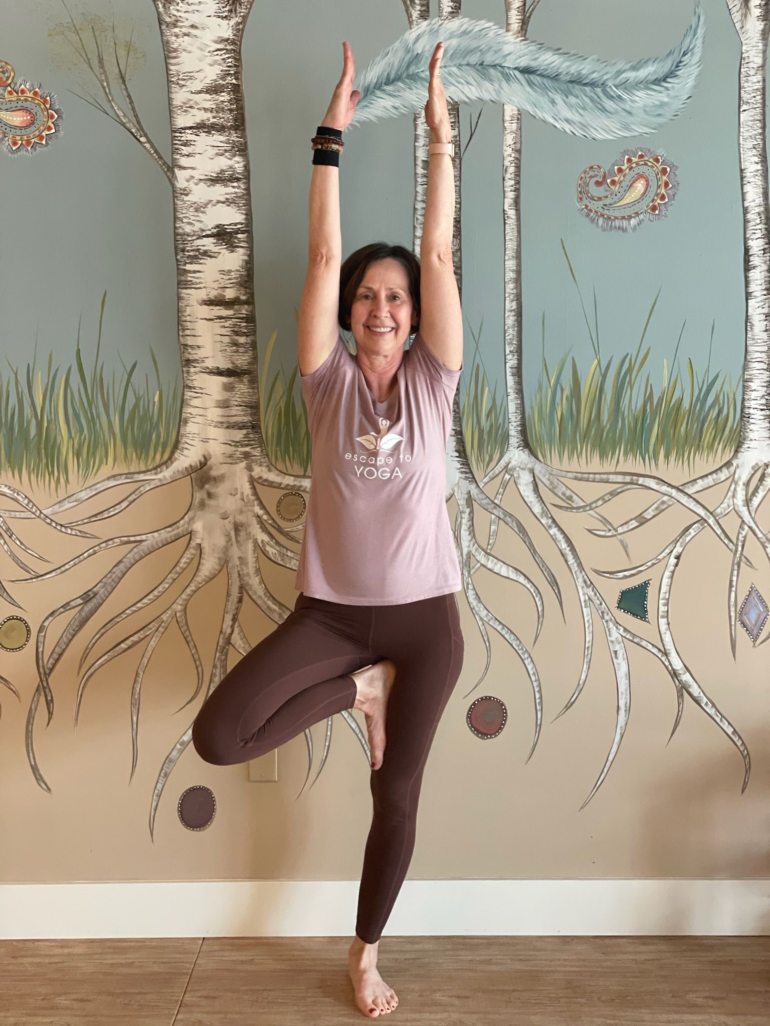 A woman is standing in a yoga pose in front of a painting of trees.