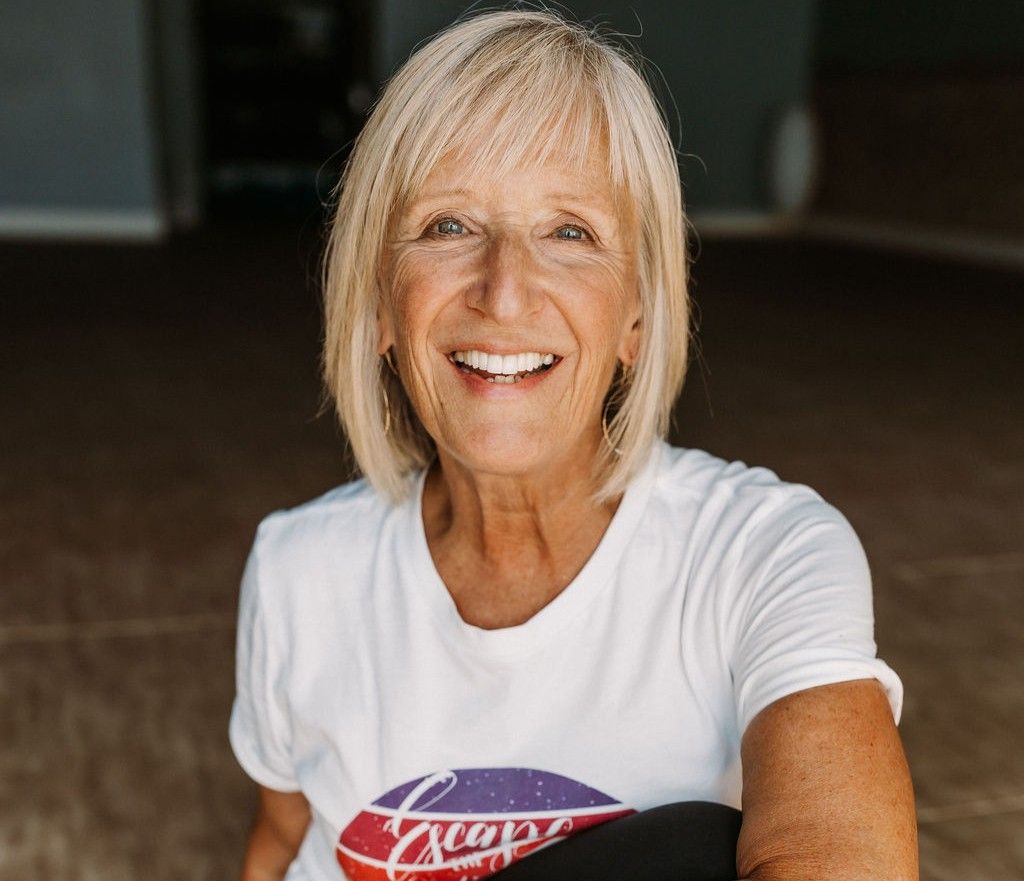 Jackie, smiling with shoulder-length blonde hair, is wearing a white t-shirt with a colorful circular logo.
