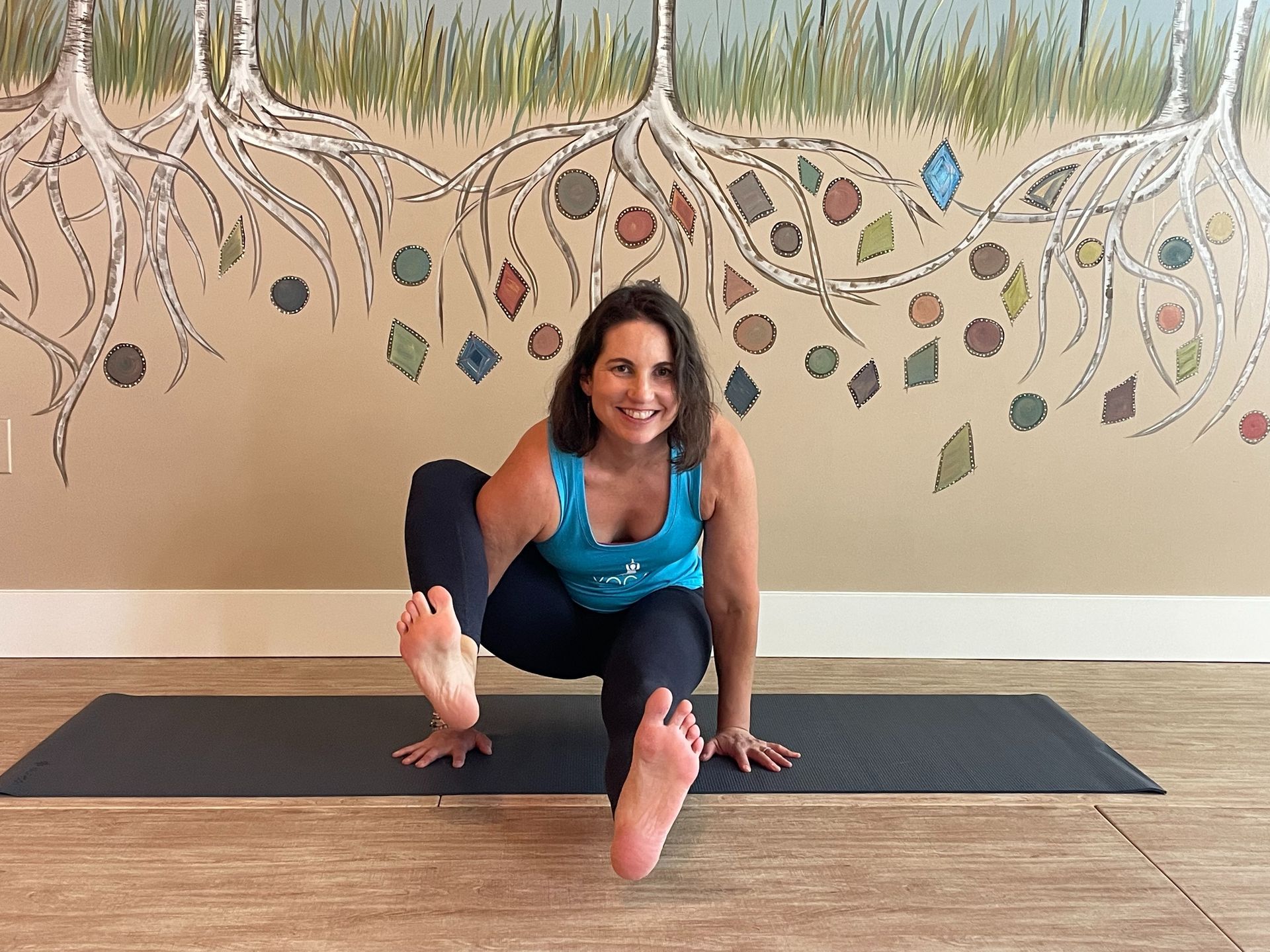 Woman smiling on a yoga mat in a seated stretch, with one foot extended toward the camera.