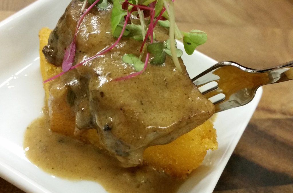 Braised meat with sauce on polenta, garnished with microgreens on a white plate