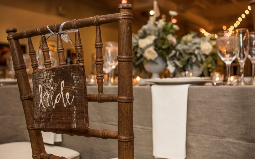 Rustic wedding reception table with a wooden chair and floral centerpiece, warm string lights in the background