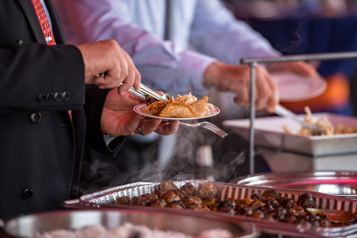 Hand serving food from a buffet line with steaming trays and utensils