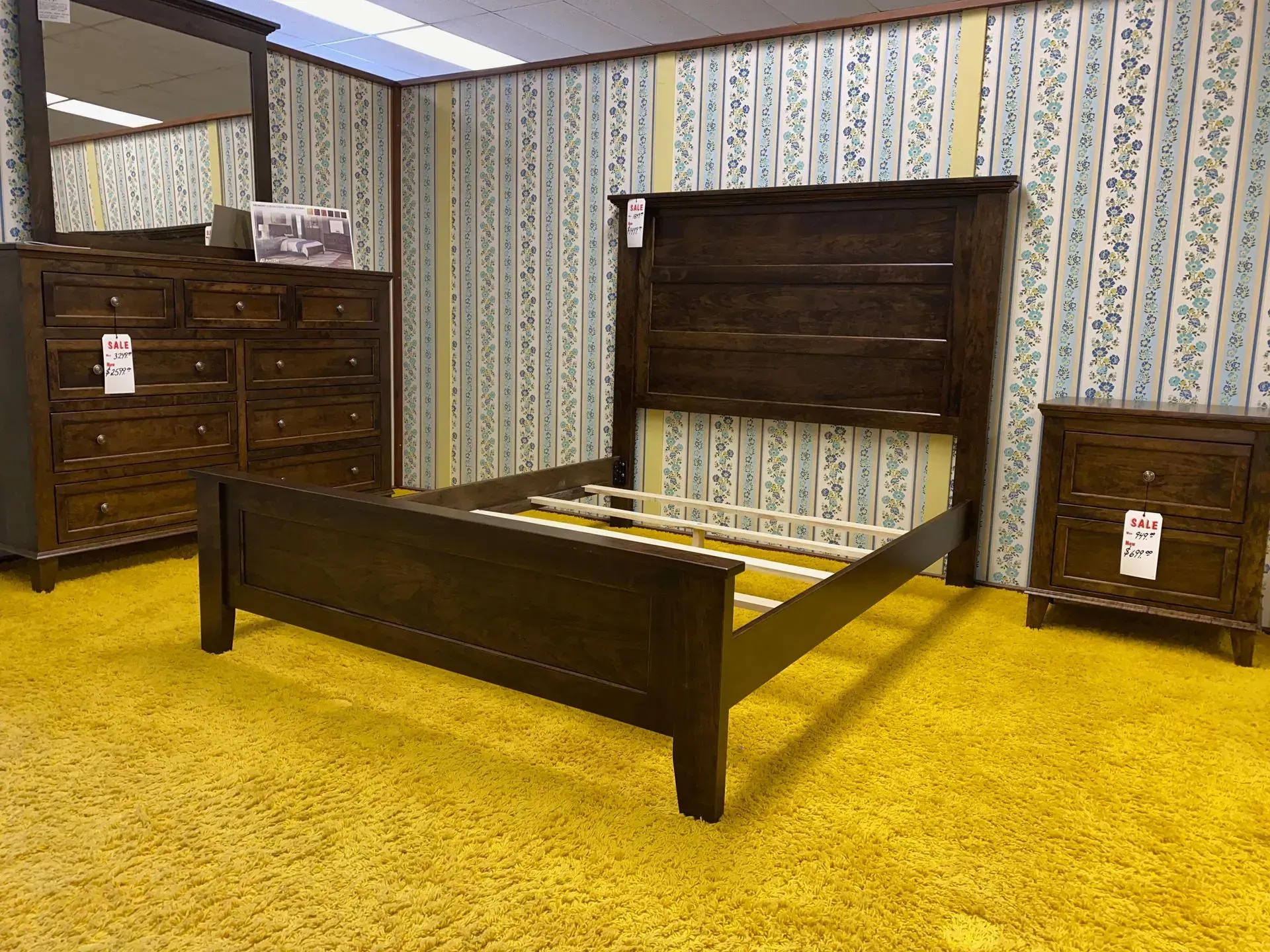 Bedroom furniture set: dark wood bed, dresser with mirror, and nightstand on yellow carpet, floral wallpaper.