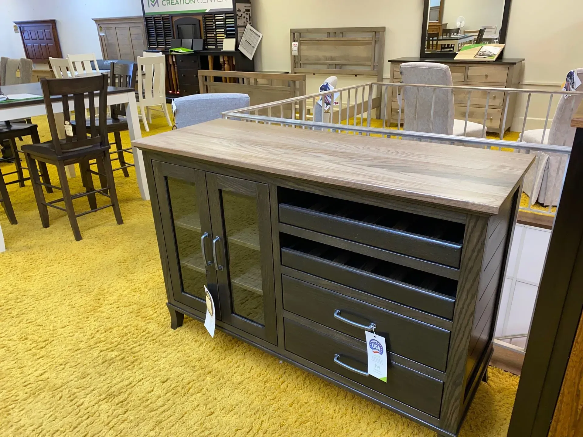 Wooden cabinet with glass doors and drawers, on a yellow carpet in a furniture store.