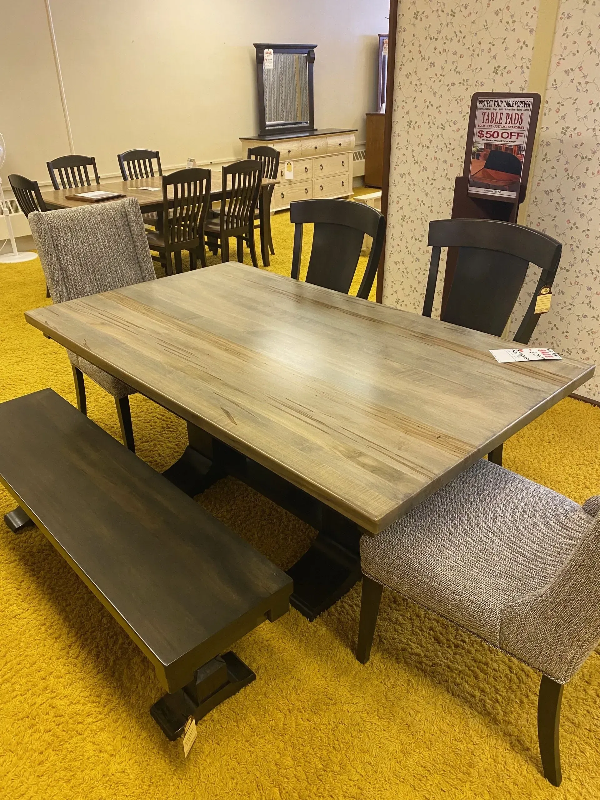 Dining table set with bench and chairs on a yellow carpet in a furniture store.