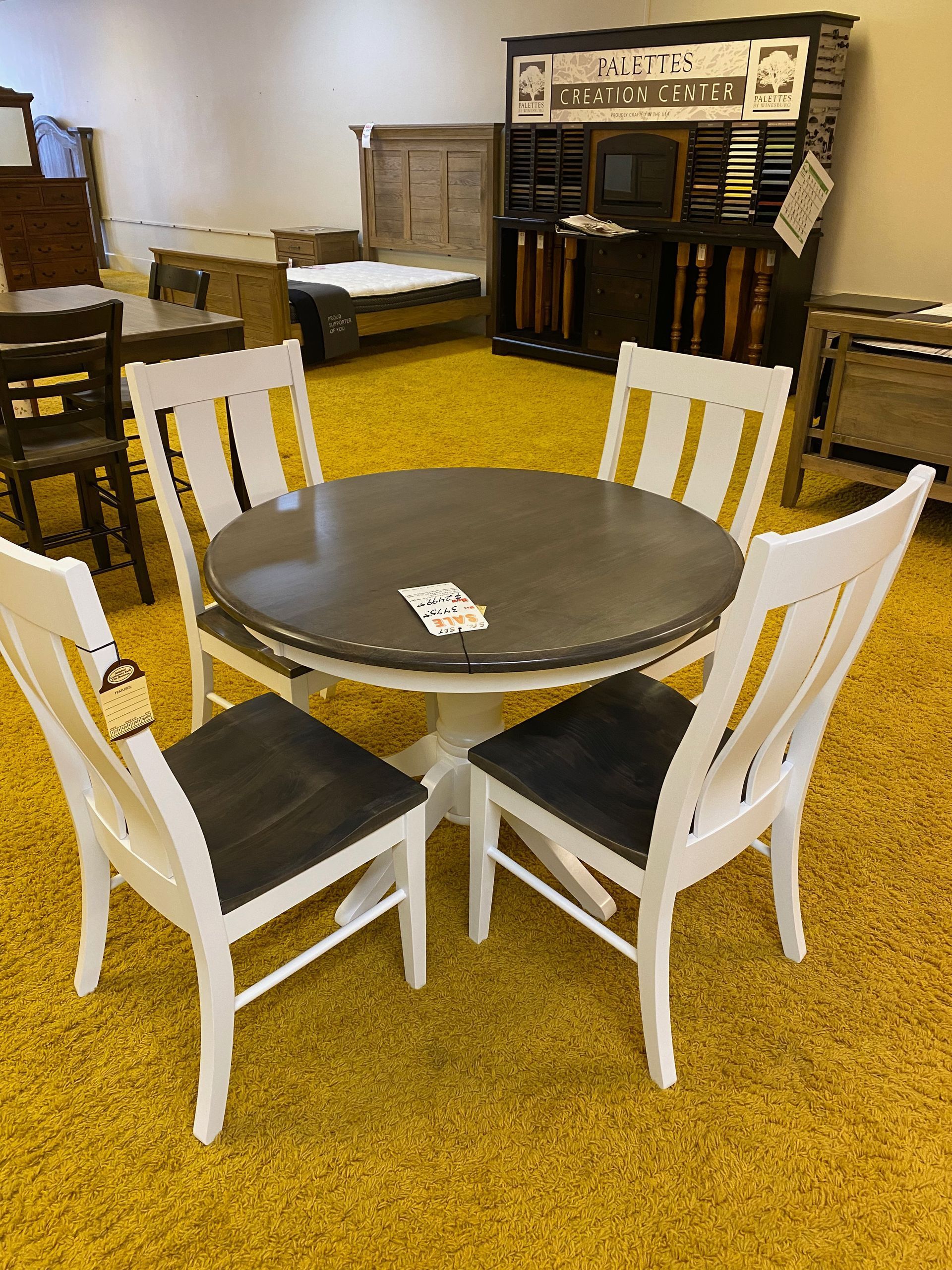 Round gray table with four white chairs, set on a yellow carpet, inside a furniture store.