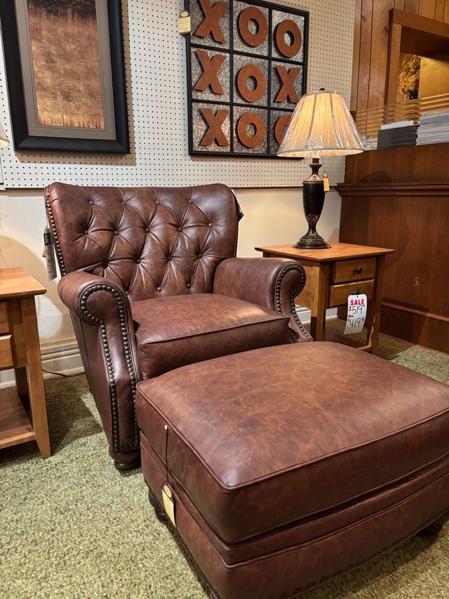 Brown leather armchair and ottoman with a button-tufted back, wooden side tables, lamp, and tic-tac-toe art in a store.