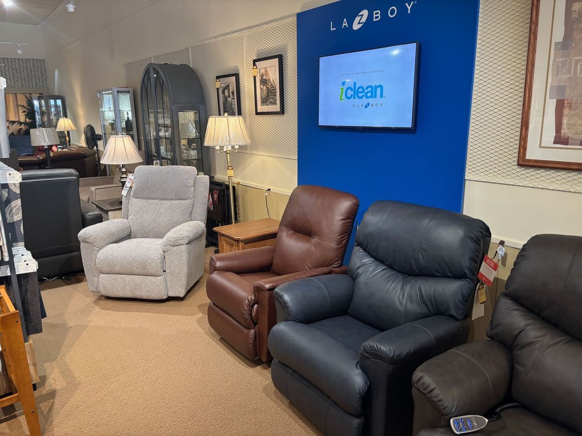 Several La-Z-Boy recliners in a showroom: gray, brown, and black leather, near a blue wall with a TV.