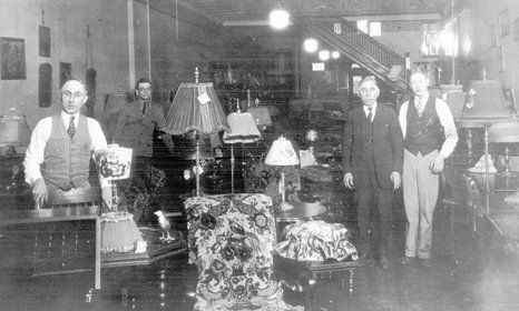 Interior of a lamp shop with several men, lamps on display, and a staircase.