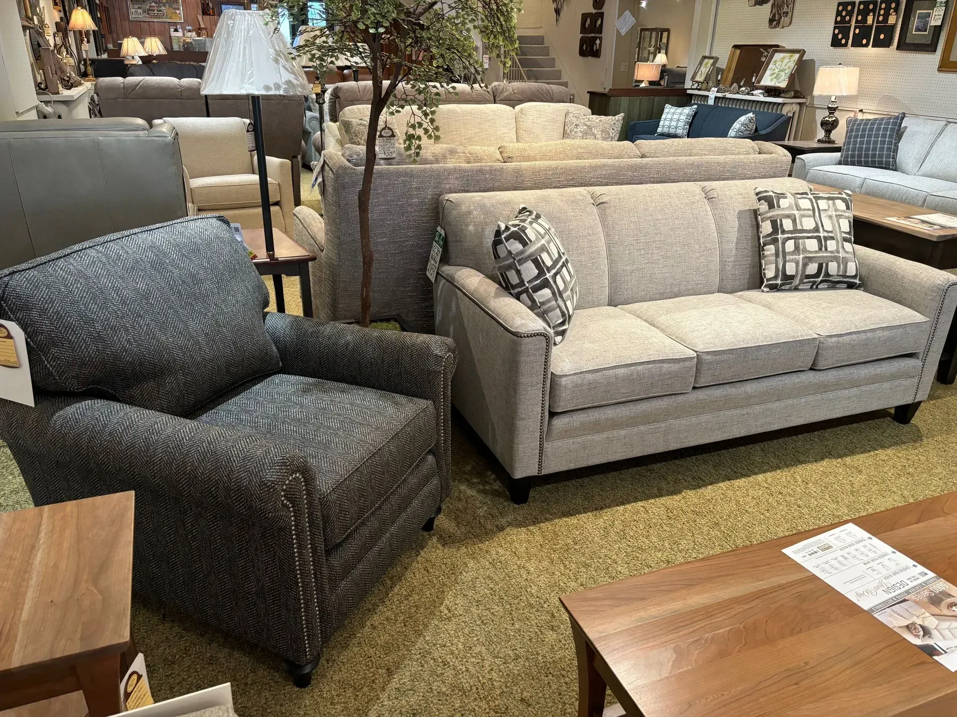 Living room furniture set in showroom: gray sofa, patterned armchair, wood tables, and neutral setting.