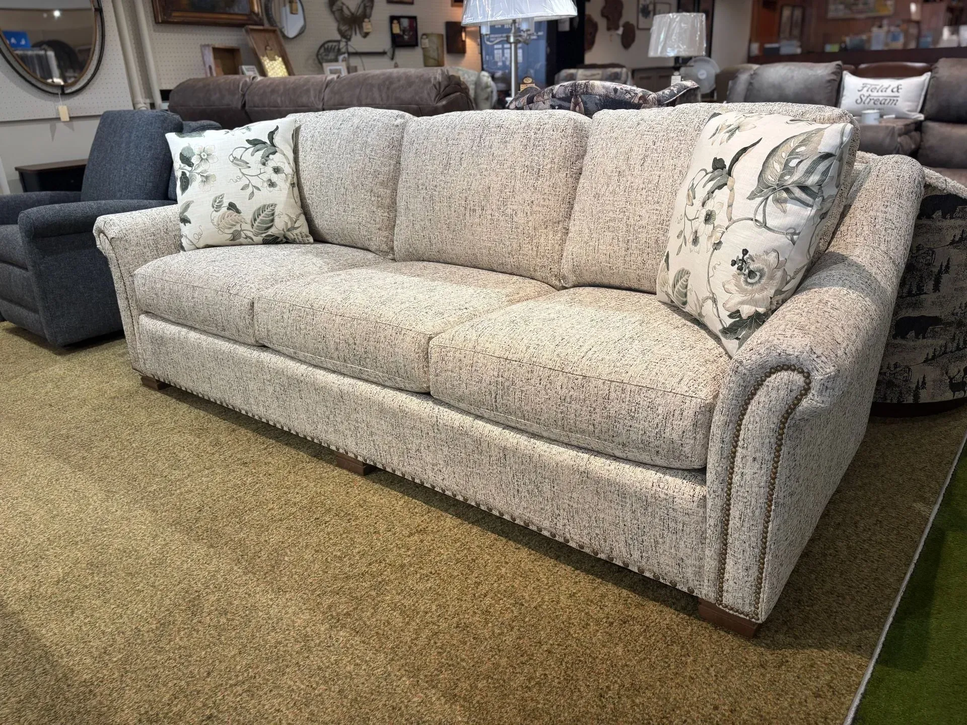 Beige speckled fabric sofa with two patterned pillows, indoors on a brown carpet.