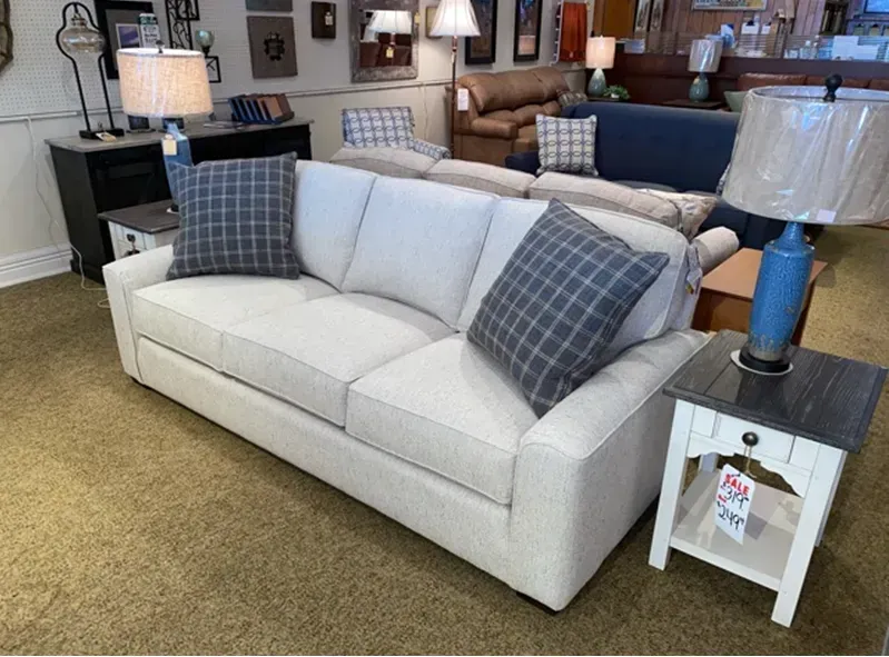 Cream-colored sofa with two plaid pillows, next to a white side table with a lamp; showroom setting.