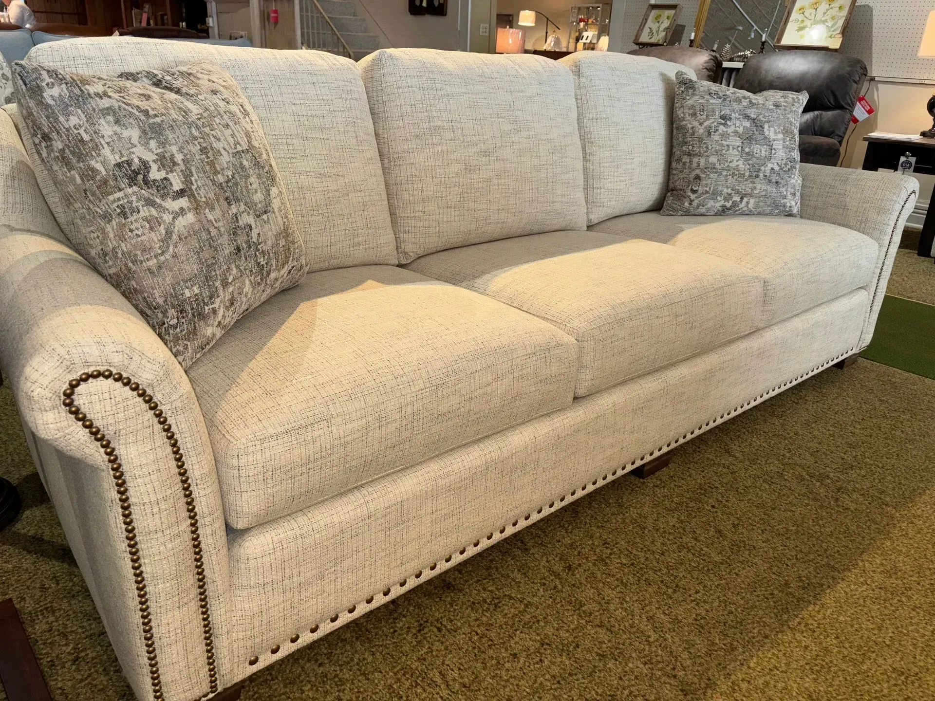 Cream-colored sofa with three cushions and two patterned throw pillows, on green carpet, in a furniture store.