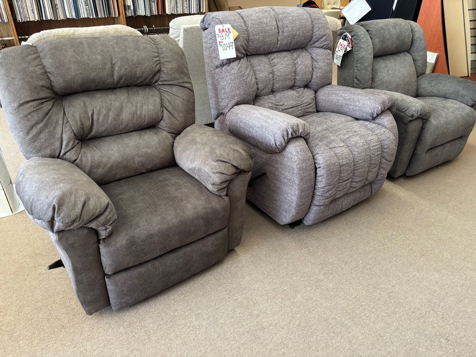 Three gray upholstered recliners on a carpeted floor, likely in a furniture store.