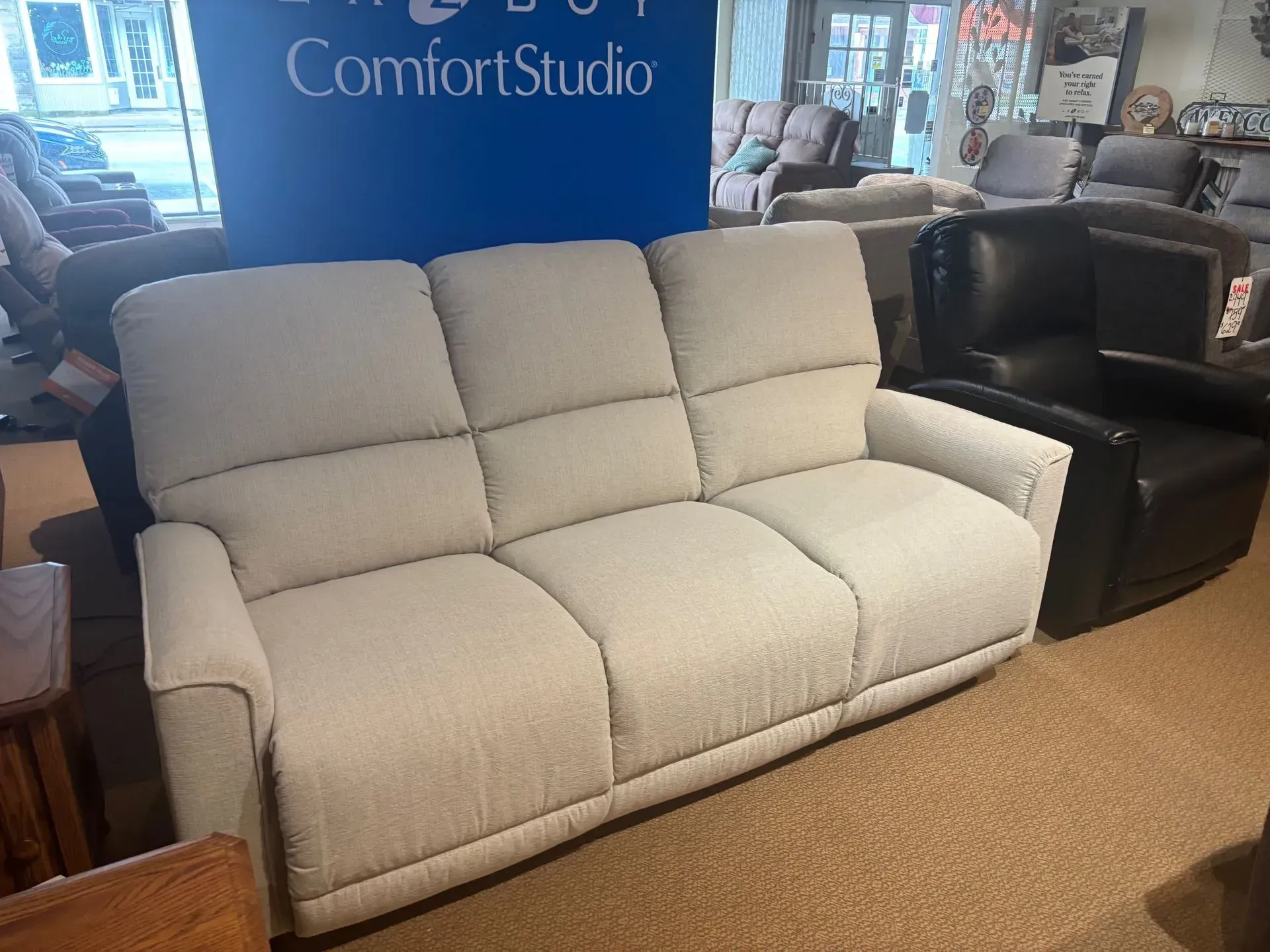 Beige three-seat reclining sofa in a furniture store. Other furniture visible in the background.
