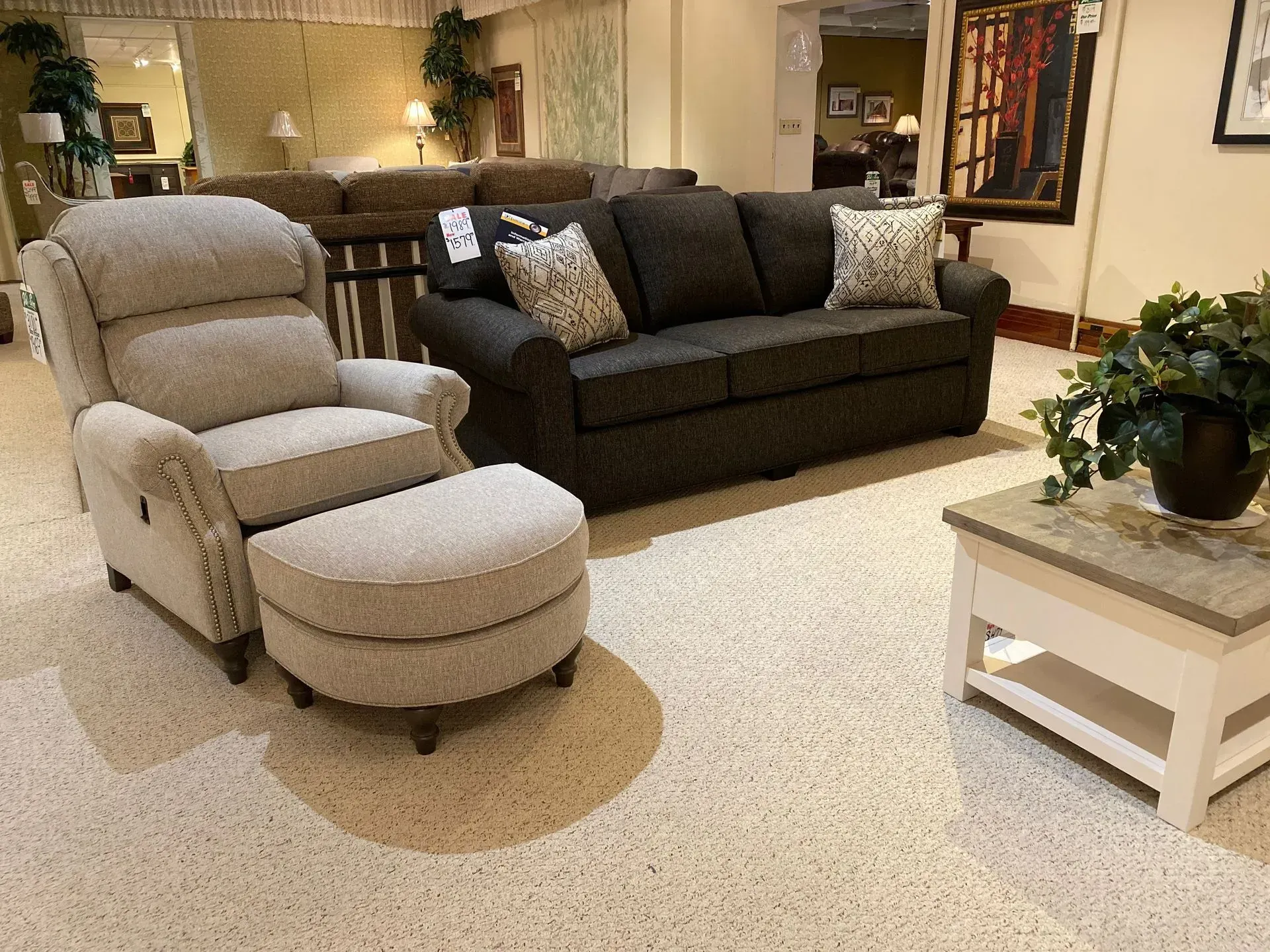 Beige recliner with matching ottoman beside a gray sofa in a showroom.