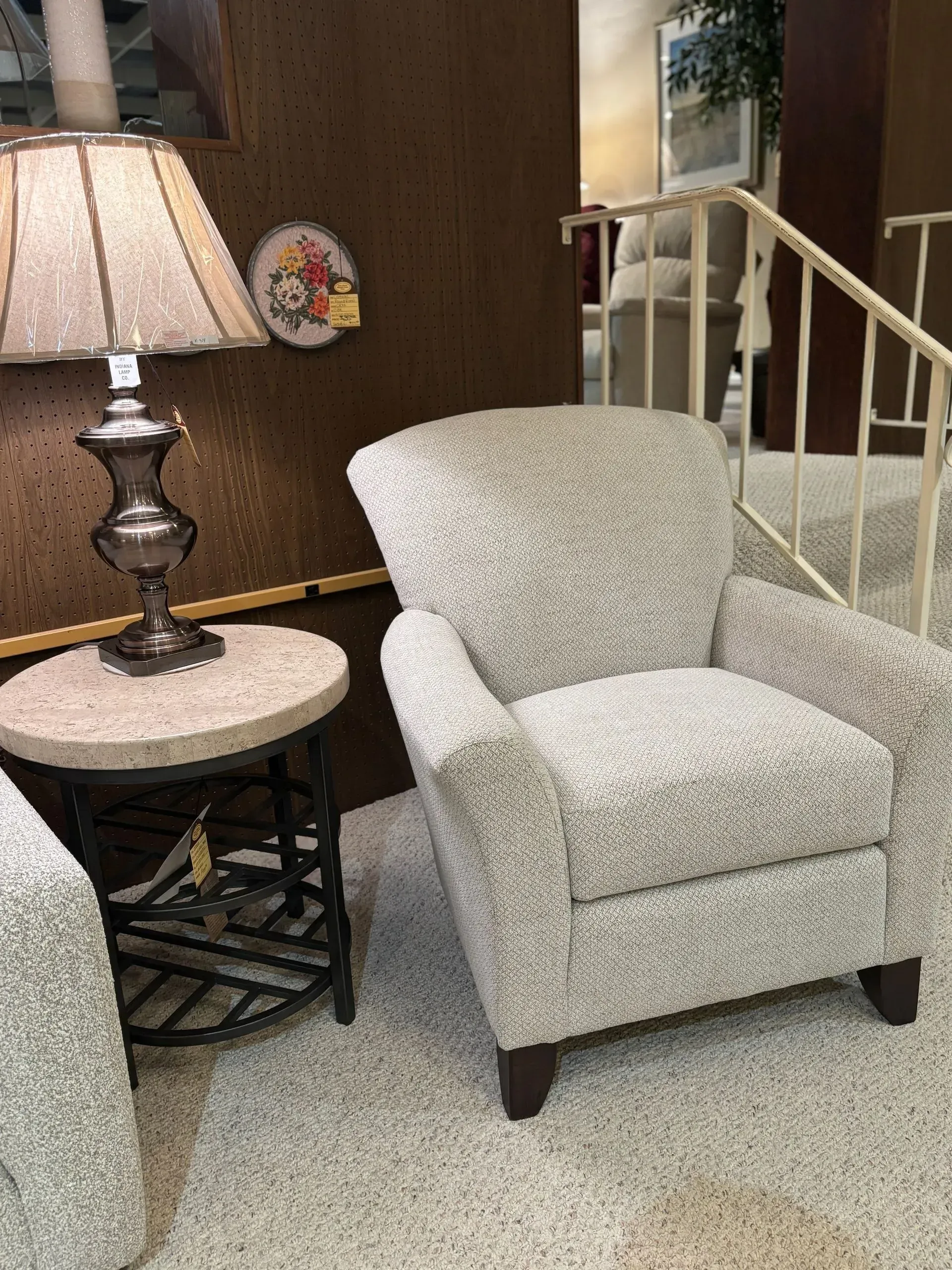 Cream-colored patterned armchair next to a round side table with a lamp, set on carpet.