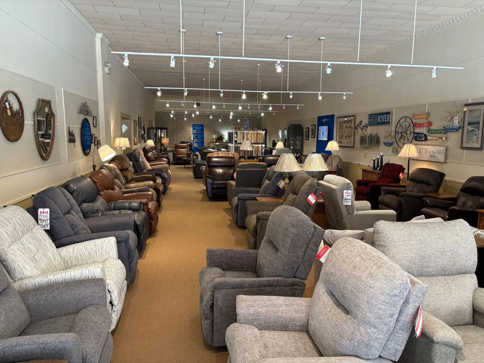 Rows of recliners in a furniture store, lit by track lighting. Beige carpet, art on the walls.