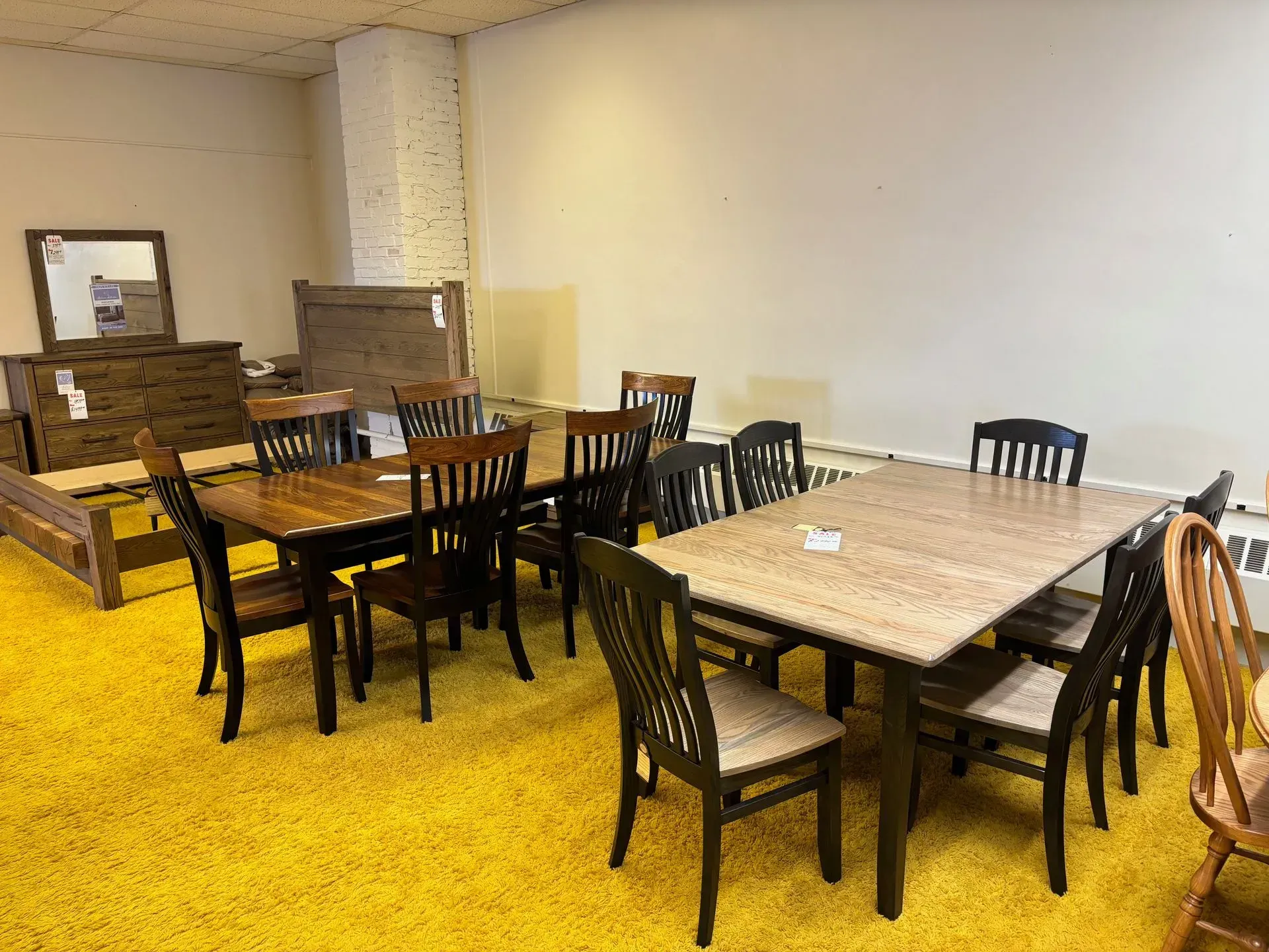 Dining room furniture display in a furniture store, with tables, chairs, and bedroom set on a yellow carpet.