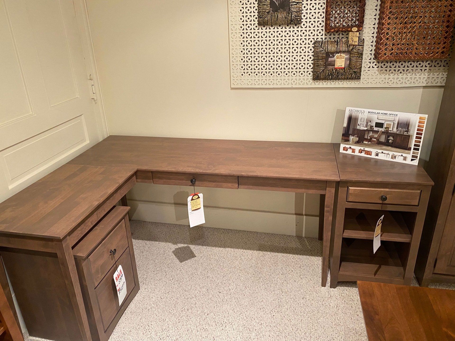 L-shaped wooden desk in a showroom with drawers and shelves; decorative wall art above.