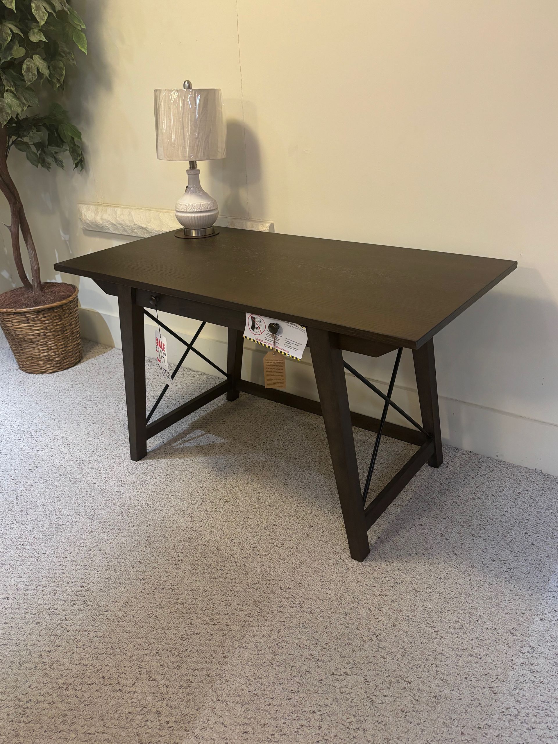 Dark brown desk with crisscross supports, a lamp, and a plant in a room with gray carpet.