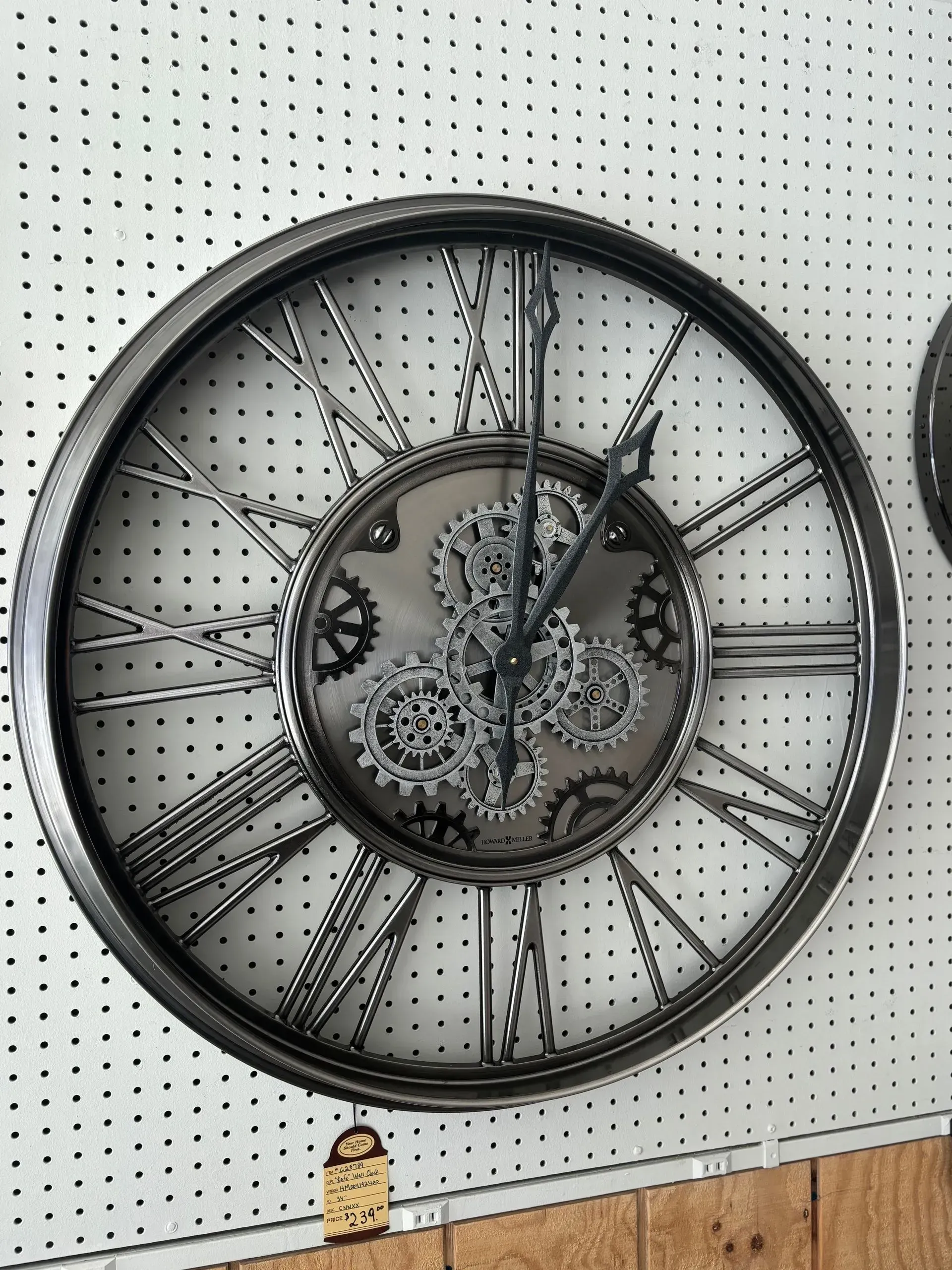 Large, round, metallic steampunk clock on white pegboard wall. Roman numerals, gear details.