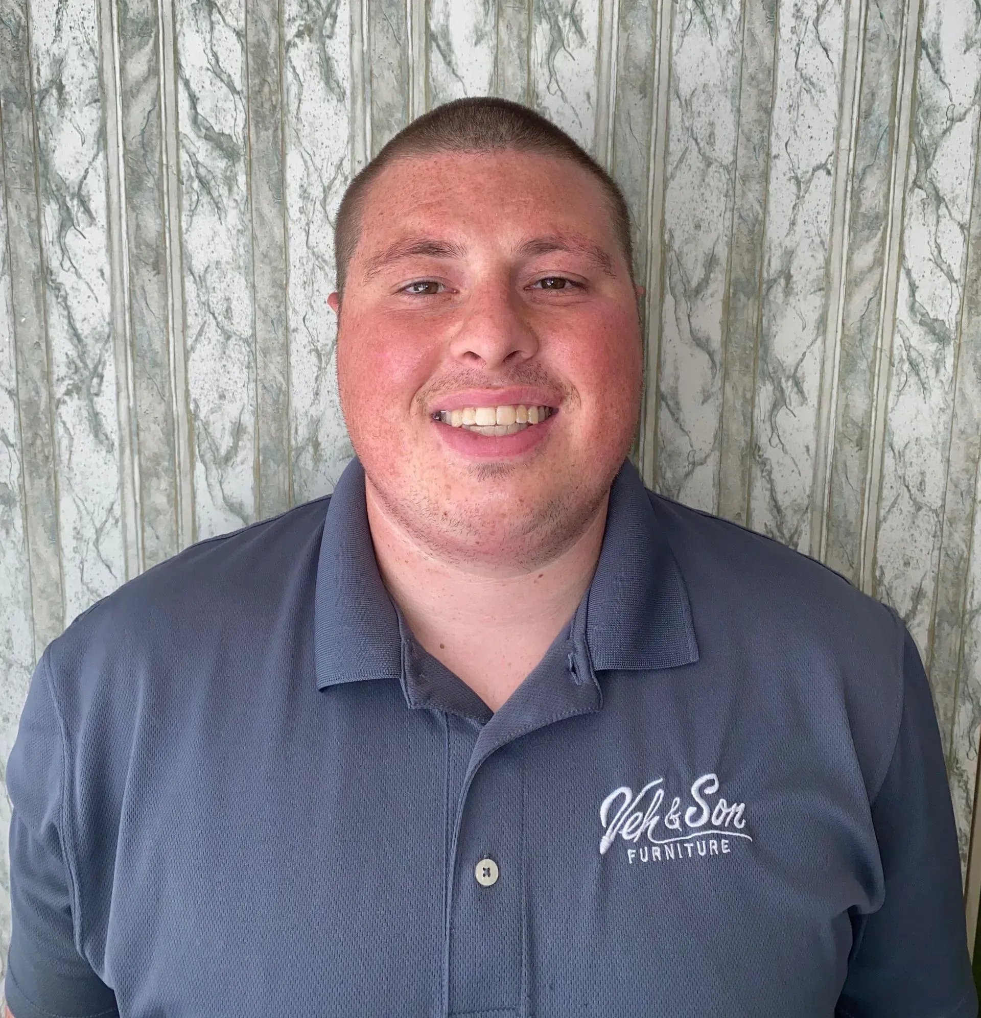 Man in a blue polo shirt smiles, posing against a patterned background. The shirt has a 