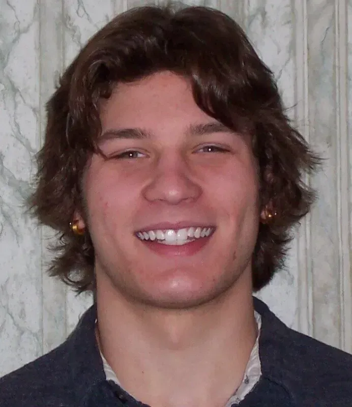 Man with brown hair and ear piercings smiles, wearing a dark shirt, indoors against a patterned background.