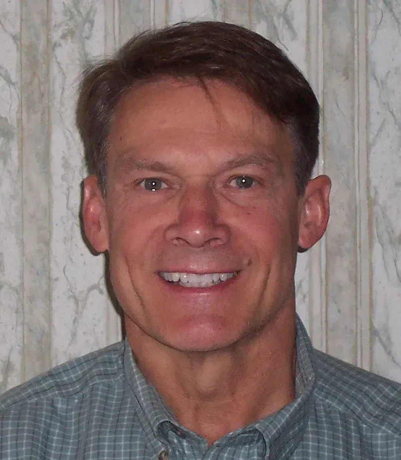 Smiling man in a green shirt; light skin, short brown hair, against a patterned background.