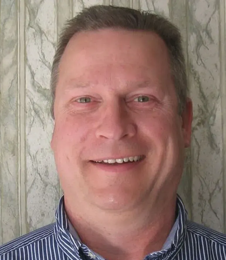 Man smiling, wearing a striped shirt, in front of a patterned wall.