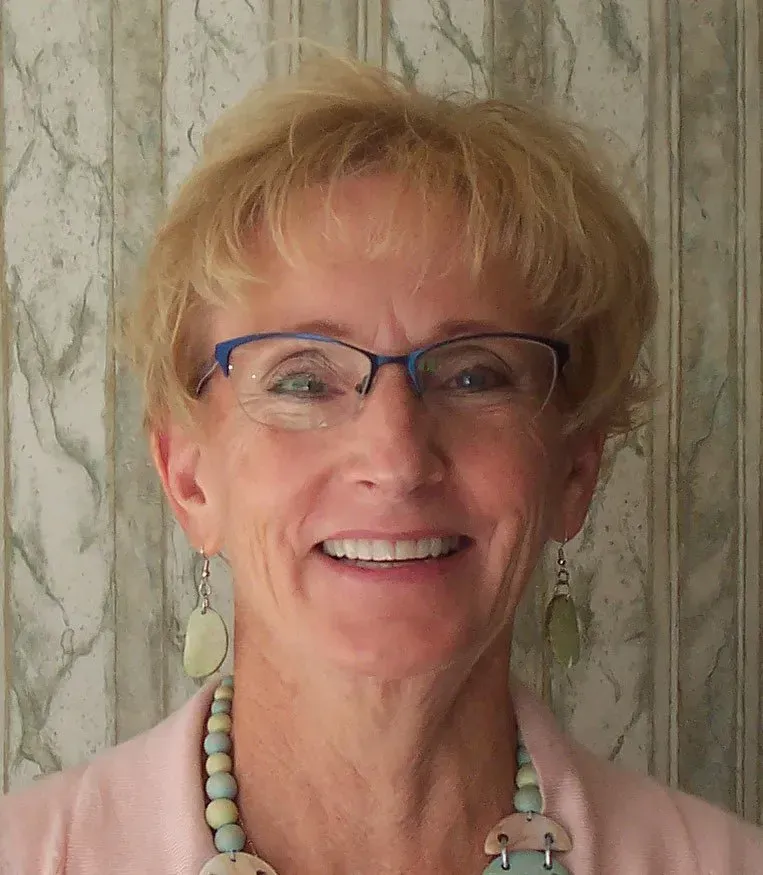 Woman with glasses smiles, wearing pink top and beaded jewelry, against a patterned background.