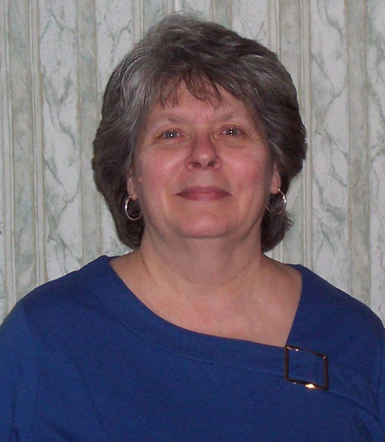 Woman with short gray hair and blue shirt, smiles in front of a striped background.