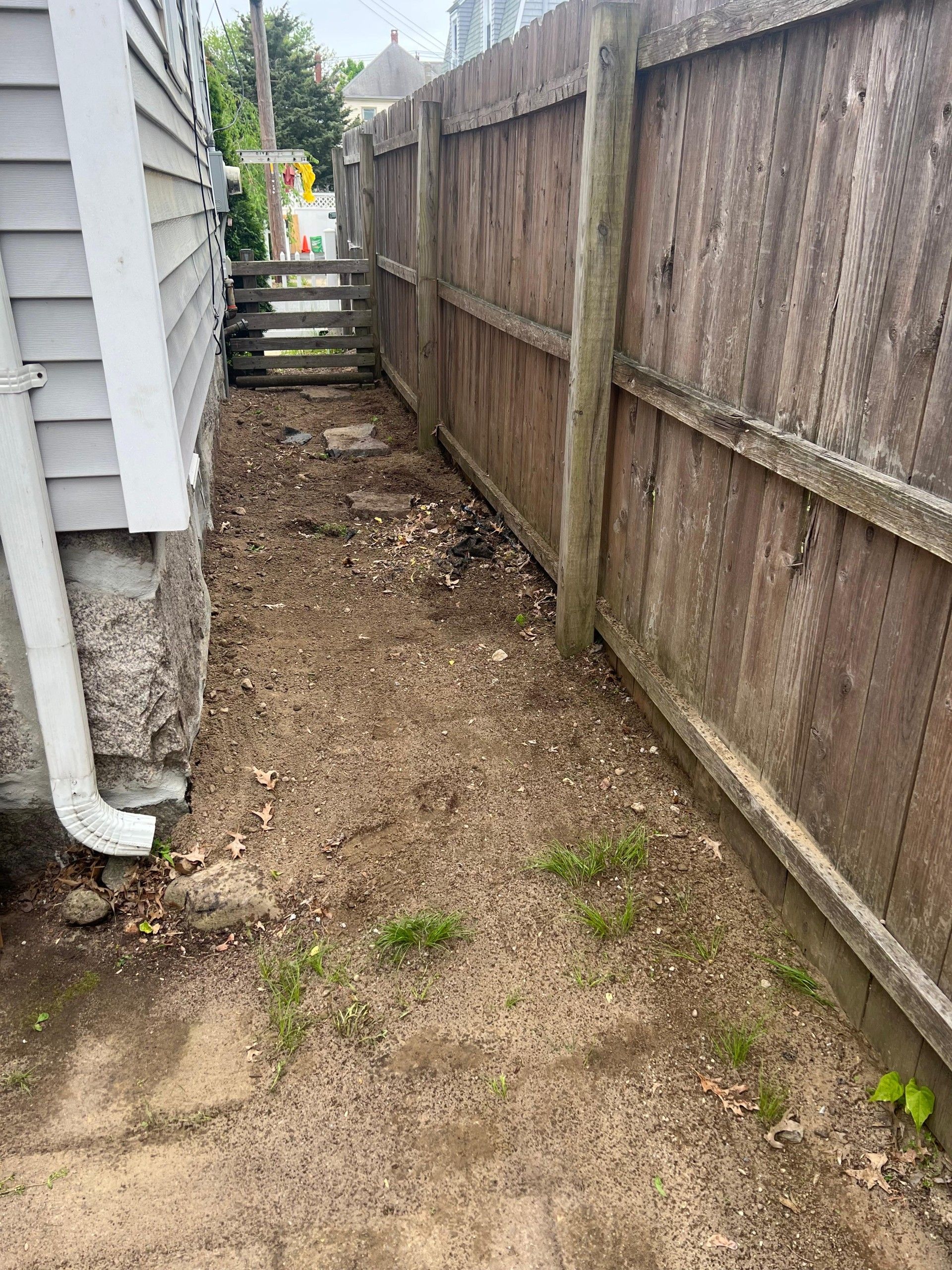Narrow dirt side yard between a house and wooden fence, with a drainpipe and steps at the far end.