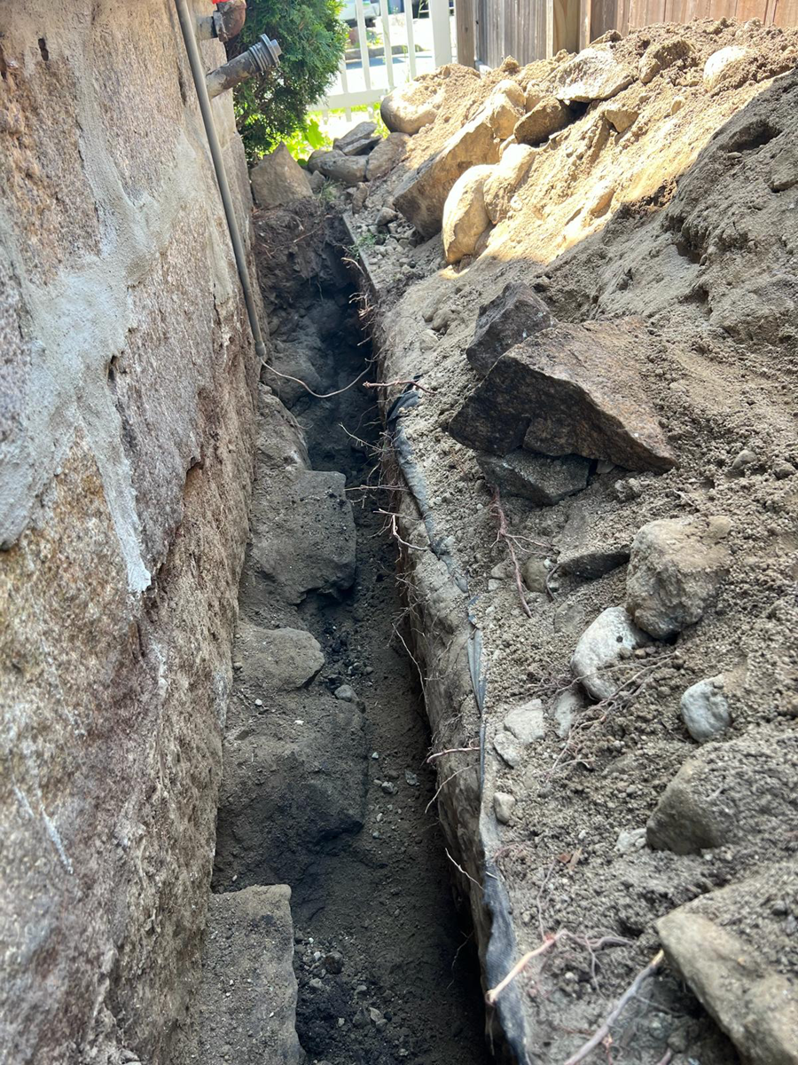 Narrow trench beside a wall, with exposed soil and debris in an urban area