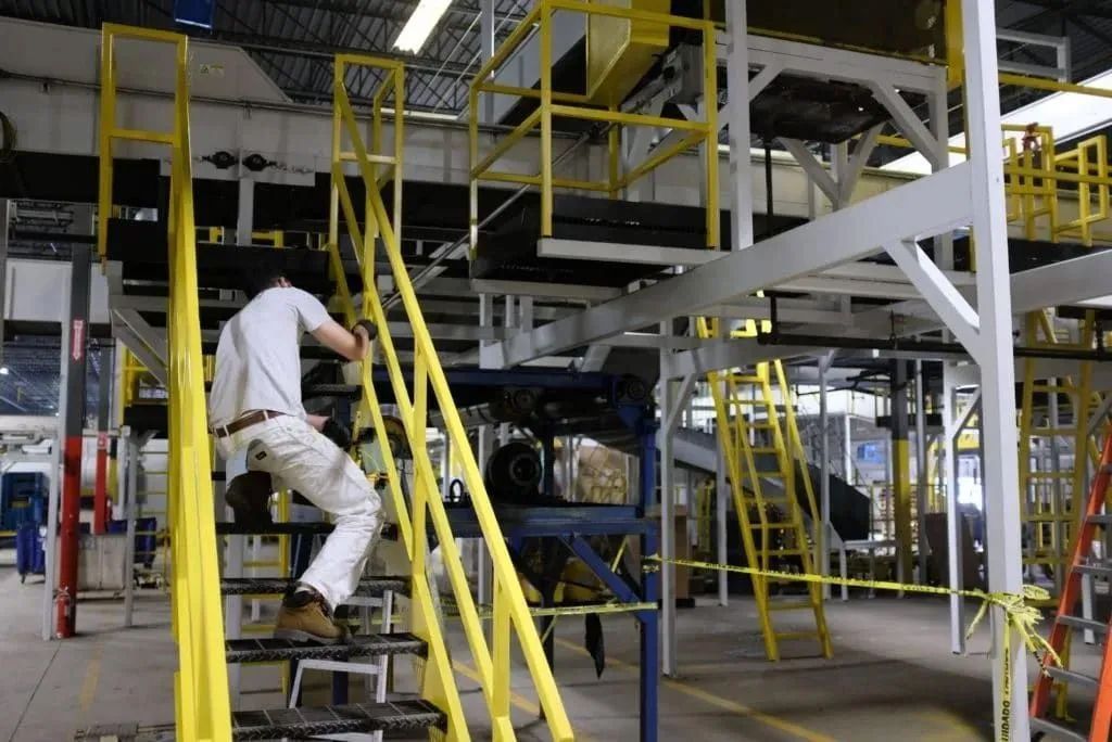 Worker climbing yellow stairs in an industrial facility with metal platforms and equipment.