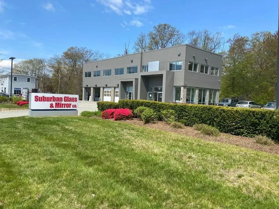 Suburban Glass & Mirror storefront with gray building, sign, shrubs, and green lawn under blue sky