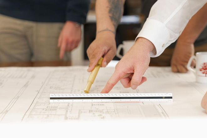 Hands measuring a floor plan with a ruler and pencil on a white drafting table