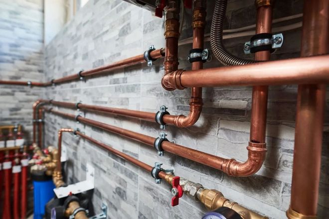 Copper plumbing pipes and valves mounted along a gray brick wall in a utility room