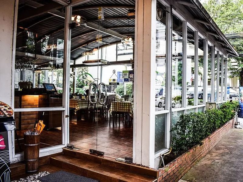 A sunlit restaurant patio enclosed in glass with checkered tables, a wooden floor, and a row of hedge plants outside.