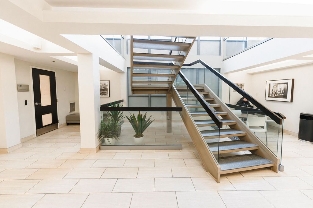 Bright modern lobby with glass-railed staircase, white tile floor, and potted plants