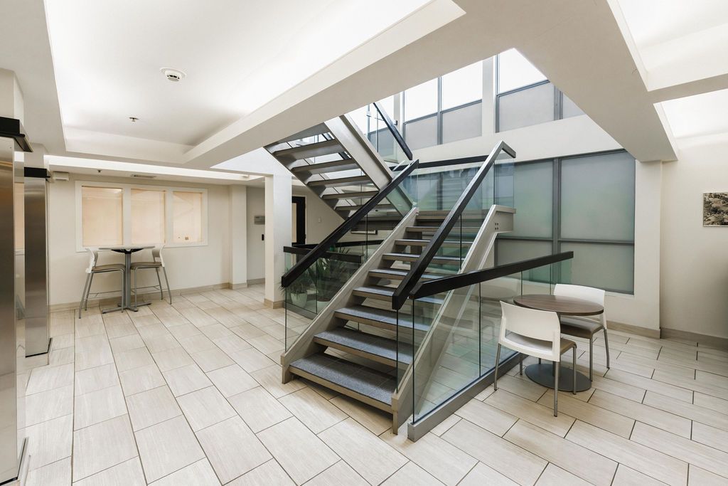 Modern lobby with central staircase, white tiled floor, glass railing, and small seating area.