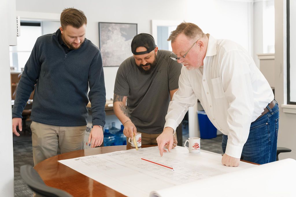 Three coworkers lean over plans on a table in a bright office, discussing a project.