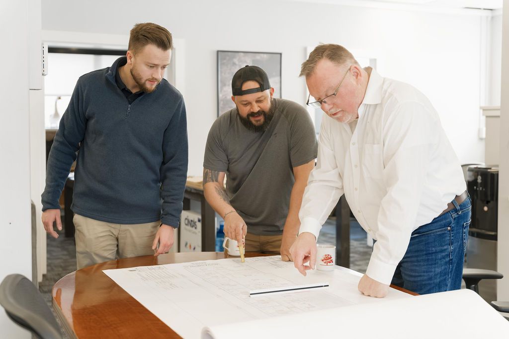 Three people leaning over a large table, reviewing a floor plan in a bright office.