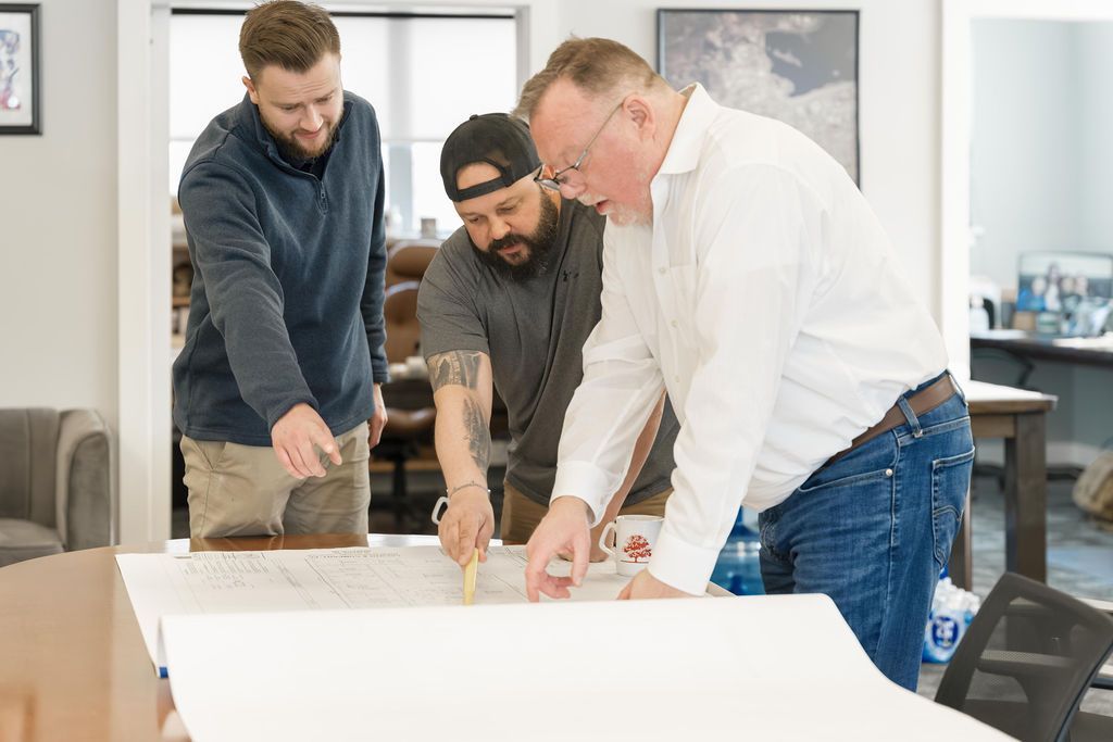 Three people collaborate around a large white board in a bright office, pointing and discussing plans.