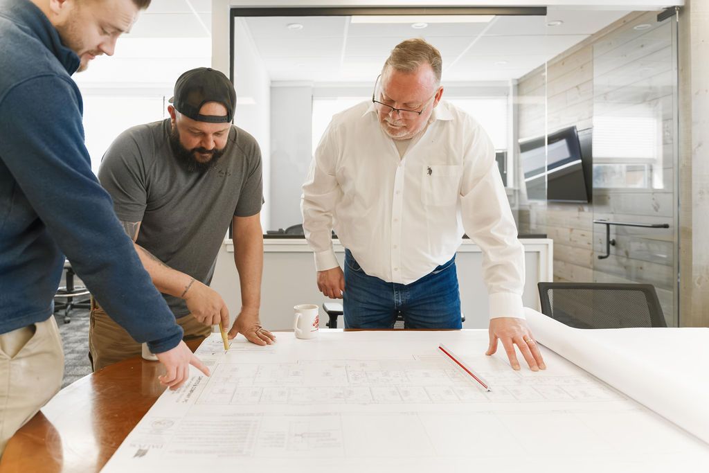 Three people review architectural plans on a table in a bright office, pointing and leaning over the drawings.