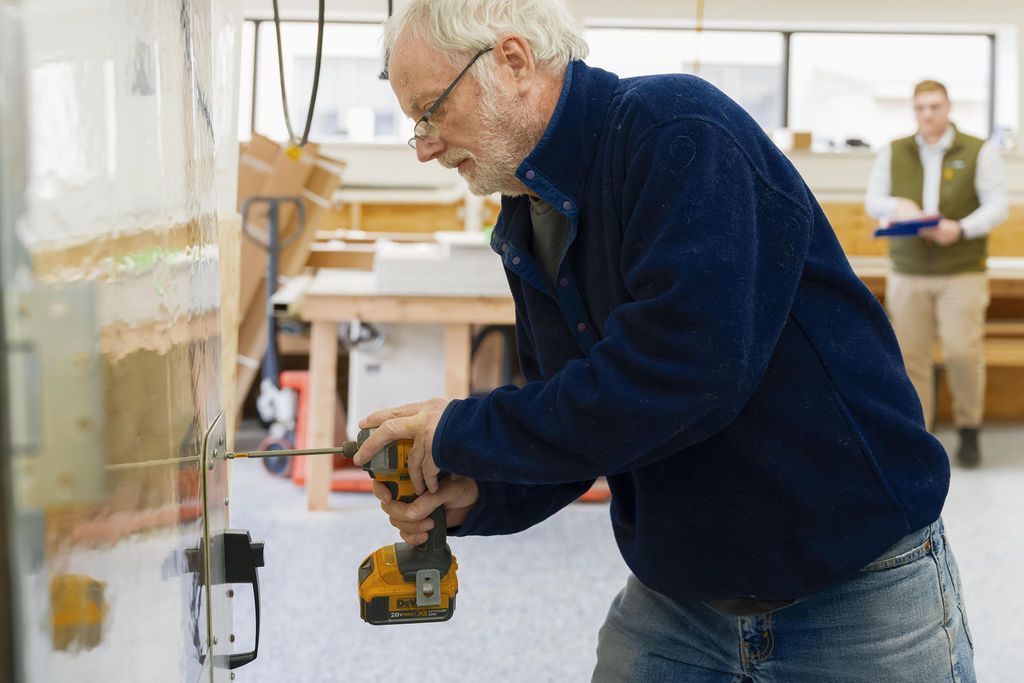 Man drilling into a wall in a bright workshop while another person stands in the background