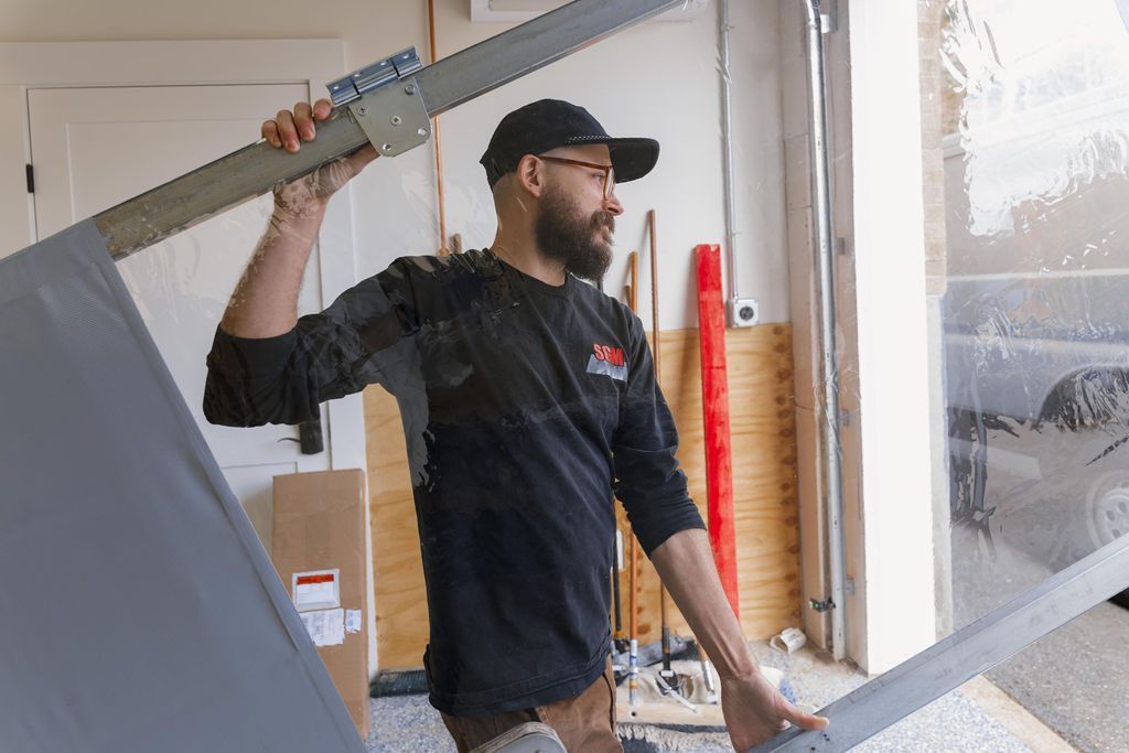 Man in black cap and jacket lifting a large window frame inside a workshop